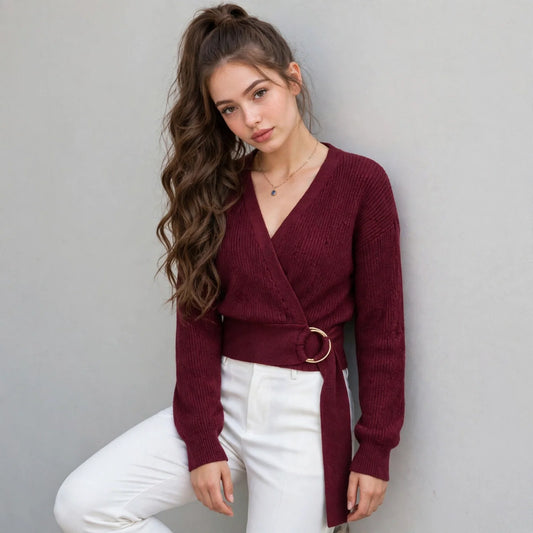 Woman modeling a burgundy wrap V-neck knit sweater with metal ring waist detail, dropped shoulders and white pants.