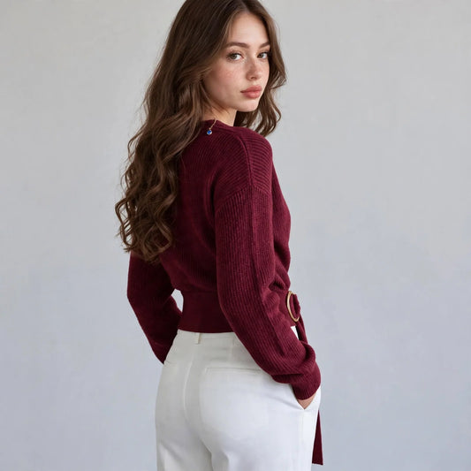 Back view of a woman wearing a burgundy wrap knit sweater with dropped shoulders and white pants, gray background.