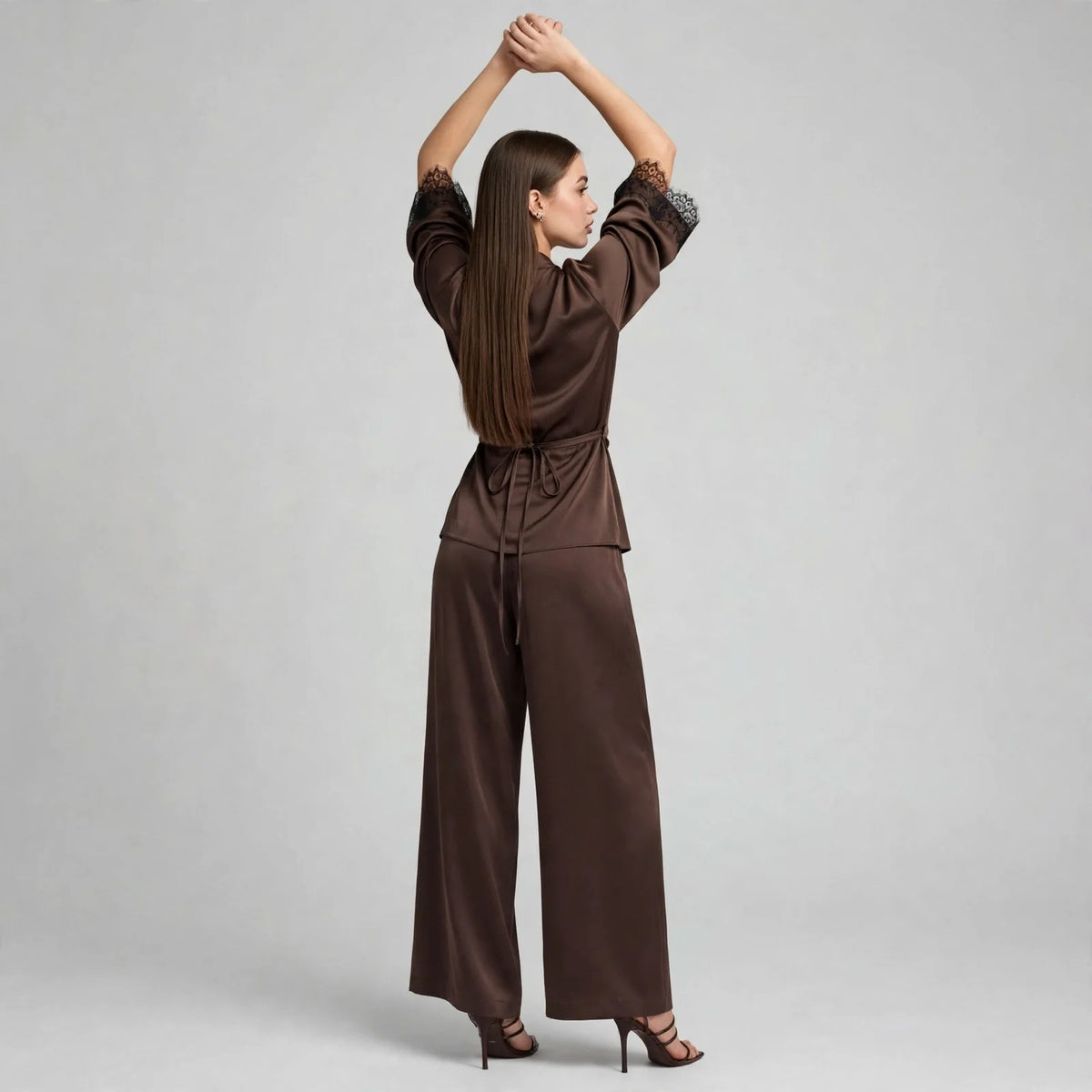 Back view of model with arms raised wearing chocolate brown satin wrap top with black lace cuffs and wide-leg pants, heels