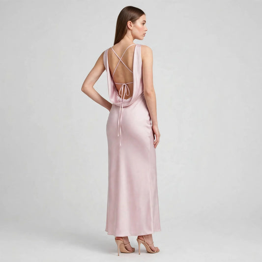 Back view of a woman in a light pink satin maxi dress with deep open back and crisscross tie straps, gray studio background.