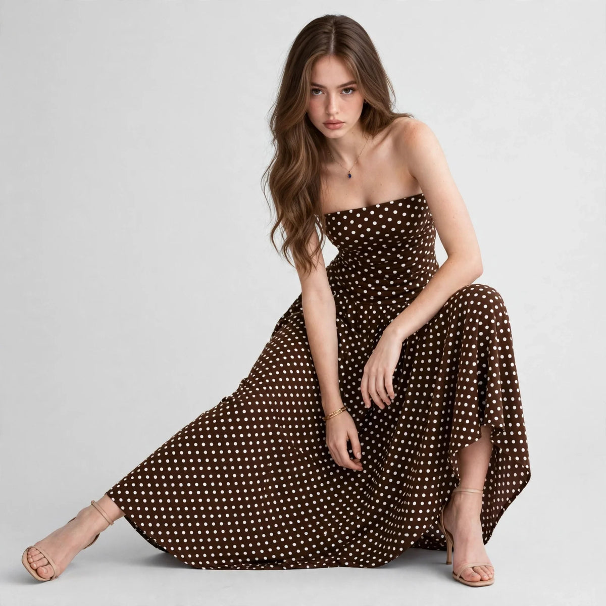 Seated model in a brown polka dot strapless maxi dress with skirt spread wide, smocked bodice, white studio background.