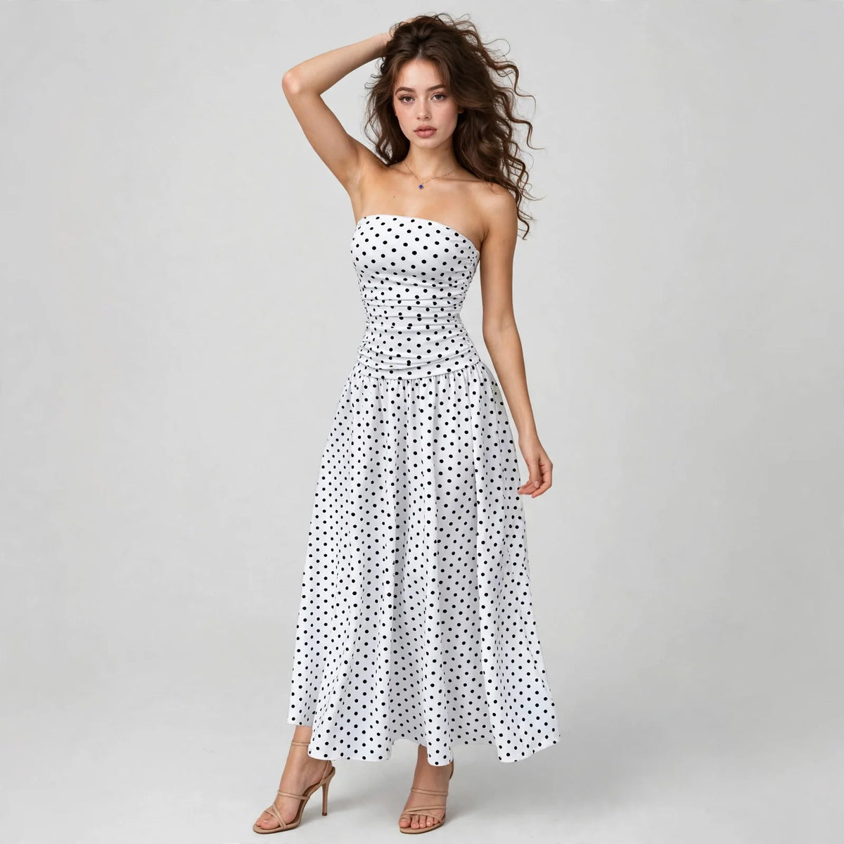 Model with hand in hair wearing a white polka dot strapless maxi dress with smocked bodice, white studio background.