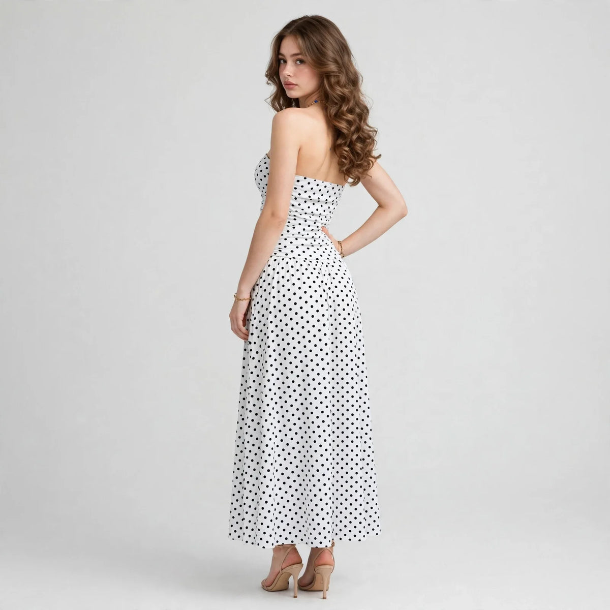 Back three-quarter view of a white polka dot strapless maxi dress with smocked bodice, hand on hip, white backdrop.