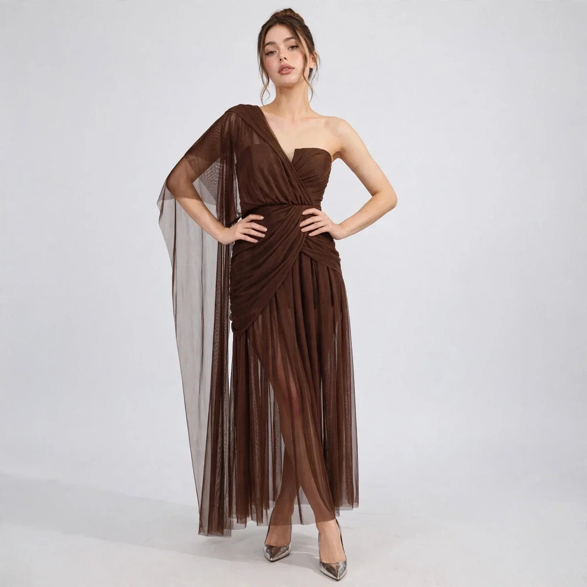 Model with hands on hips wearing brown one-shoulder mesh dress with ruched bodice and flowing skirt, front view