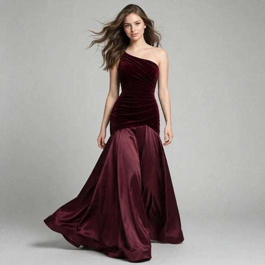 Woman mid-stride in a burgundy one-shoulder floor-length dress with ruched bodice and flowing skirt, gray studio backdrop.