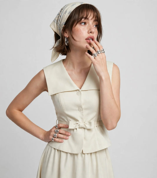 Close-up of a model in a cream sleeveless collared top with tie belt, wearing a headscarf, gray studio background.