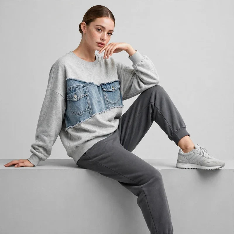 Woman seated wearing a gray denim-patchwork sweatshirt with dark joggers and white sneakers, light gray background.