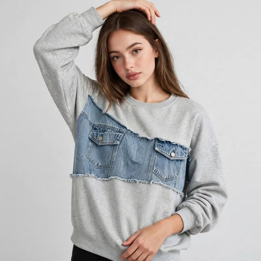 Woman with a hand in her hair wearing a gray dropped-shoulder sweatshirt with a denim patchwork yoke, gray background.