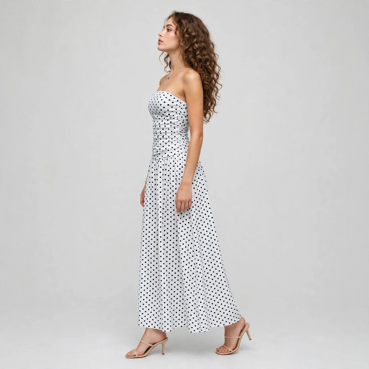 Side profile of a white polka dot strapless maxi dress with smocked bodice and flowing skirt, white studio backdrop.