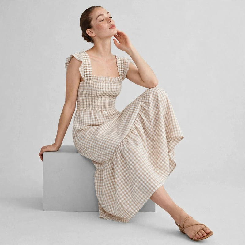 Model seated on grey block in beige gingham plaid maxi dress with ruffle sleeves and tiered skirt, side view studio shot.