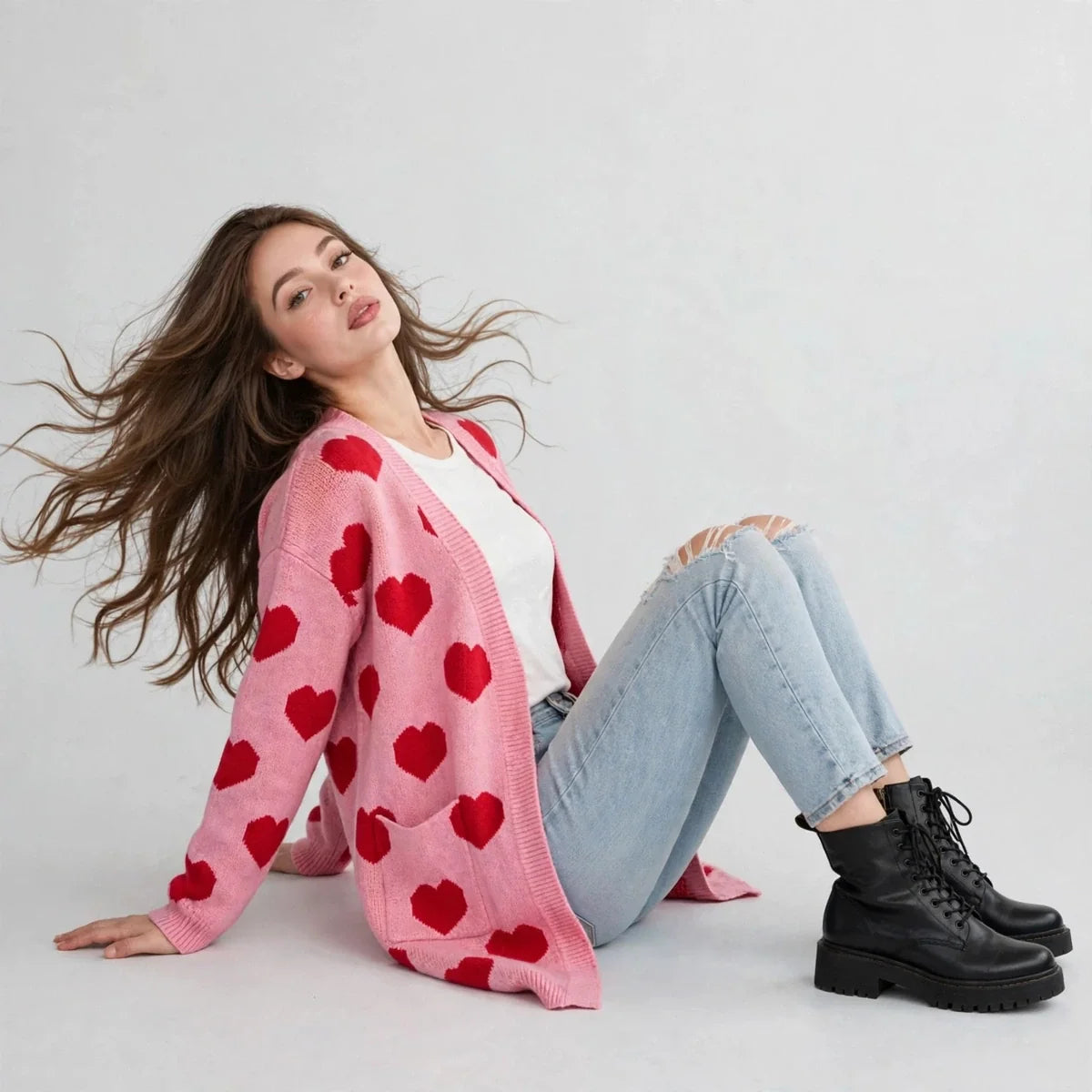 Model reclining in a pink heart-print cardigan over a white top with ripped jeans and black boots, gray background.