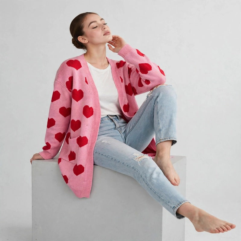 Model seated barefoot on a block in a pink cardigan with red hearts over a white top and ripped jeans, gray background.