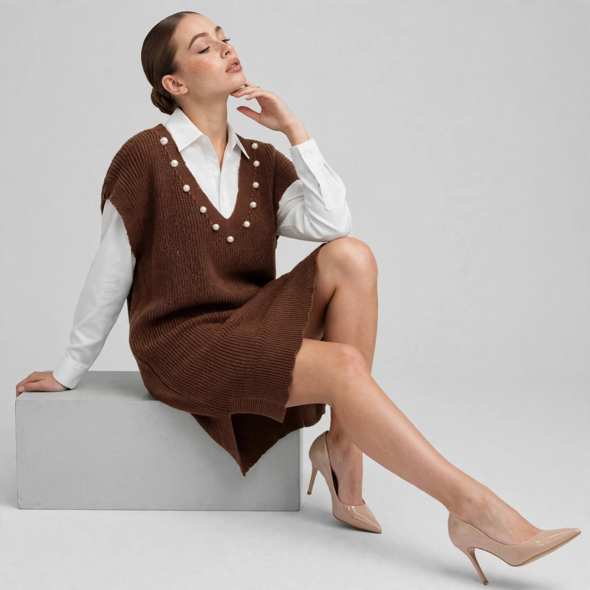 Woman seated with a hand near her chin in a brown pearl-embellished V-neck sweater dress and beige heels, gray background.