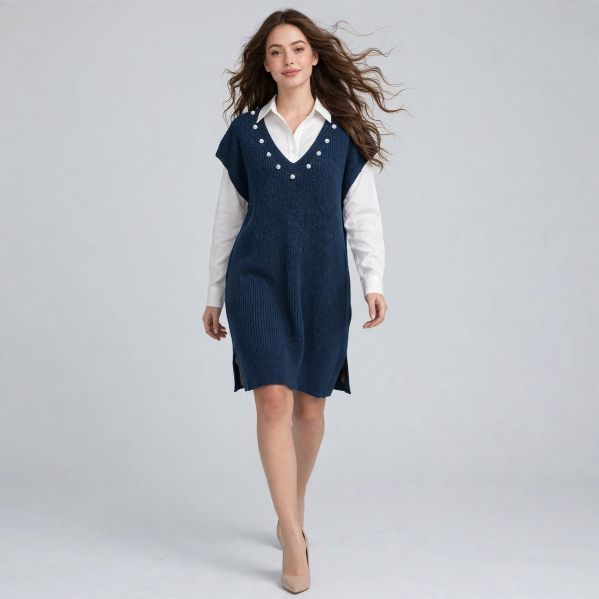 Woman walking in a navy sleeveless V-neck sweater dress with pearl embellishments over a white shirt, gray background.