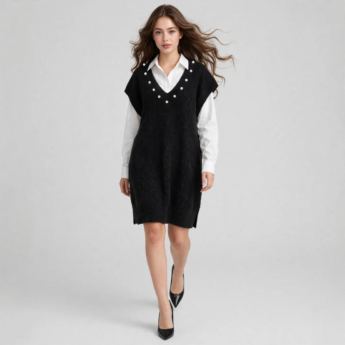 Woman walking in a black sleeveless V-neck sweater dress with pearl embellishments and black heels, gray background.