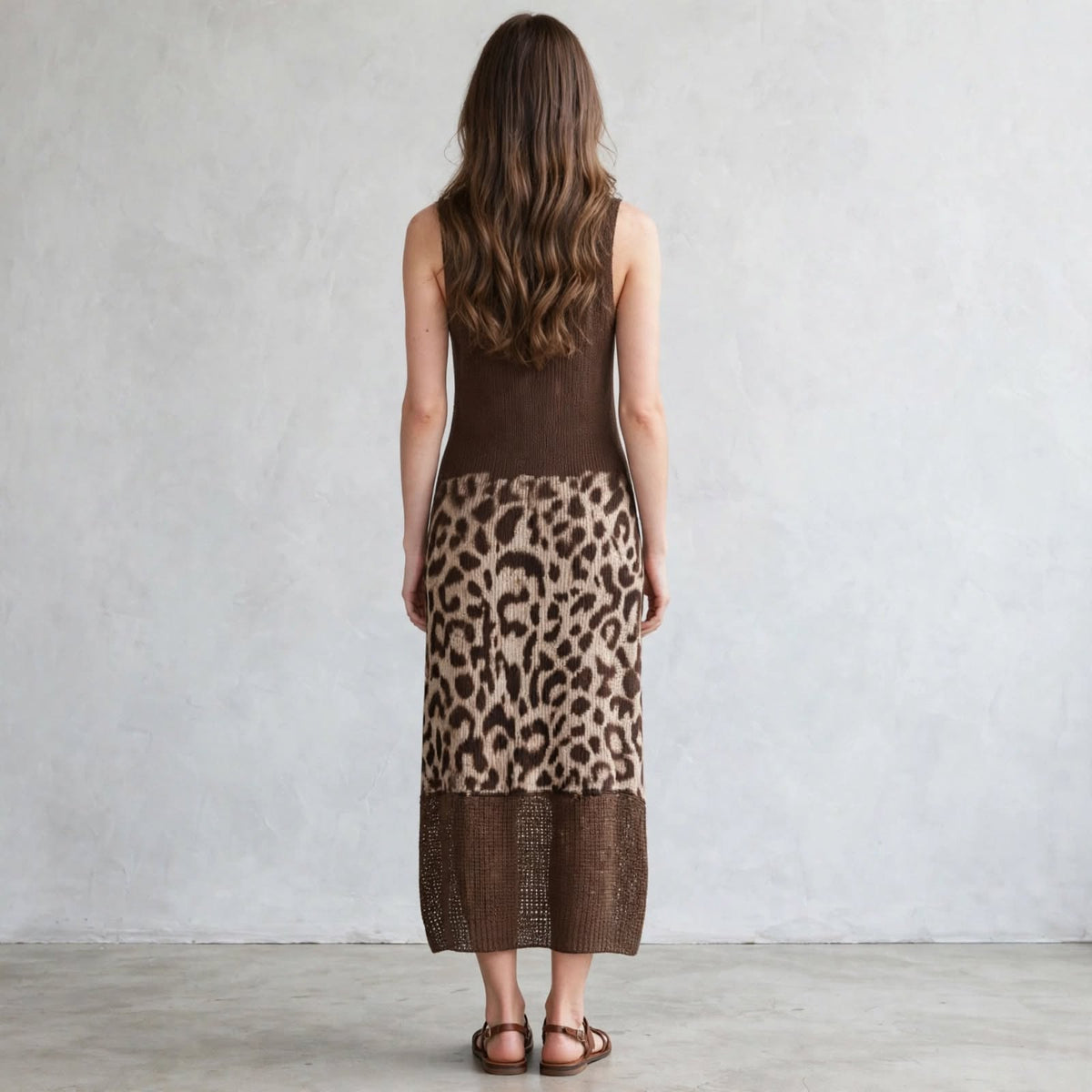 Back view of a brown sleeveless knit midi dress with leopard color-block panel on the skirt, model standing, gray studio.