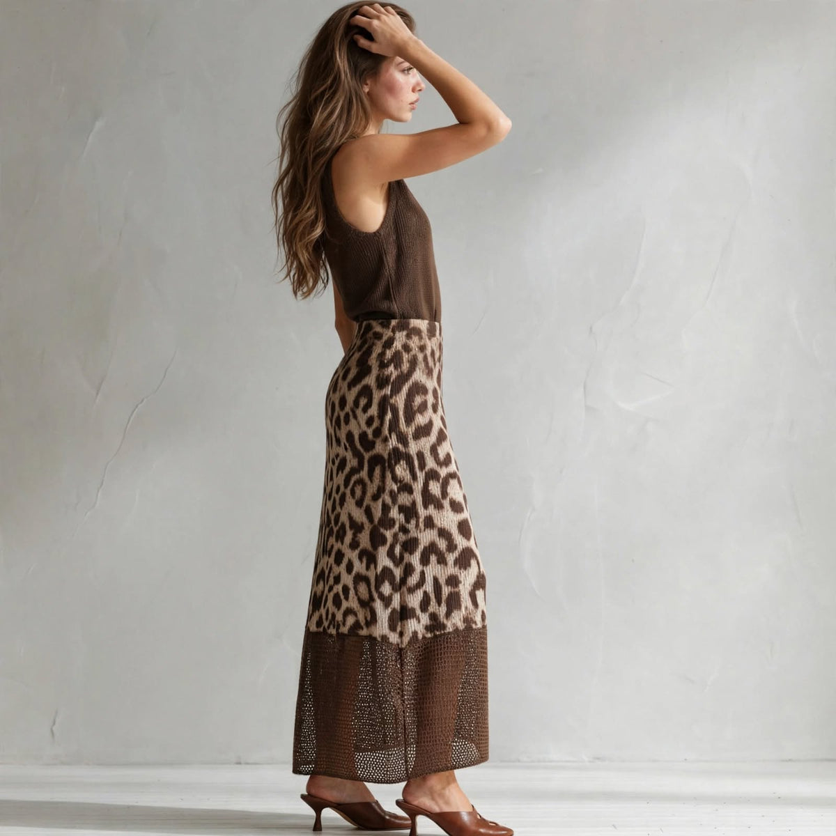 Side view of a brown sleeveless sweater dress with leopard color-block skirt panel, model with hand in hair, gray studio.
