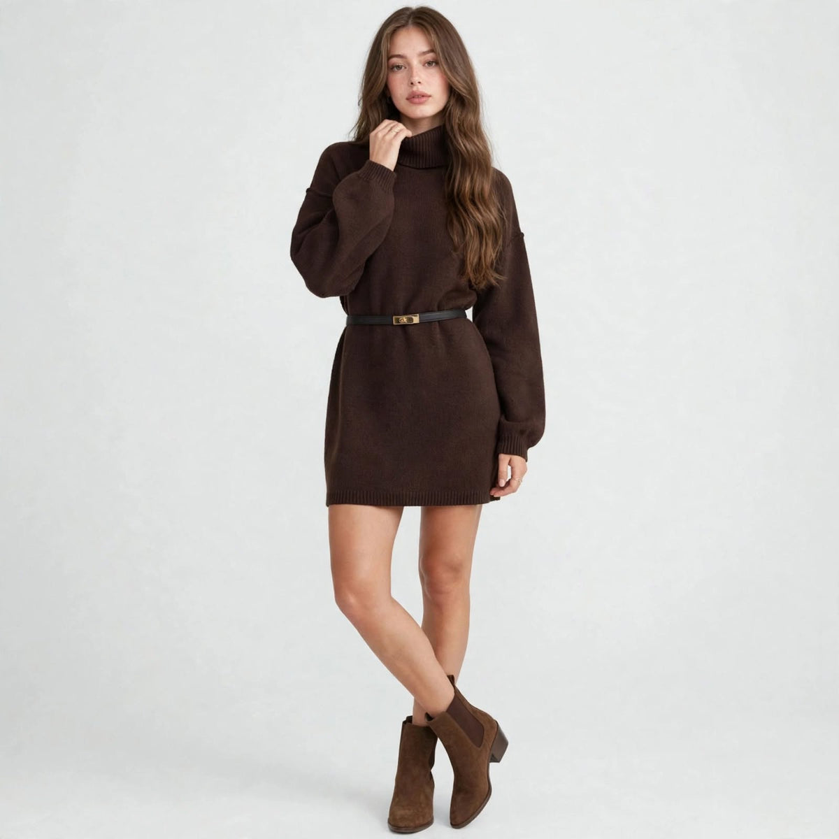 Woman modeling a brown turtleneck sweater mini dress with thin black belt, hand at chin, brown boots, gray studio background.