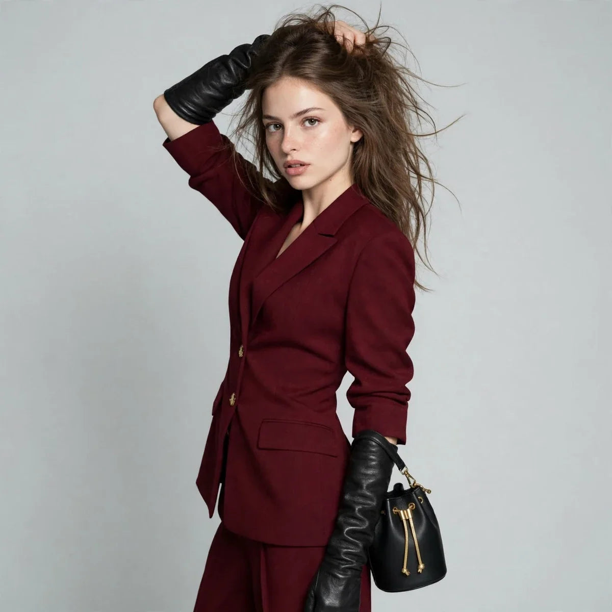 Woman with a hand in her hair wearing a burgundy blazer pants set, black gloves, and a black handbag, gray background.