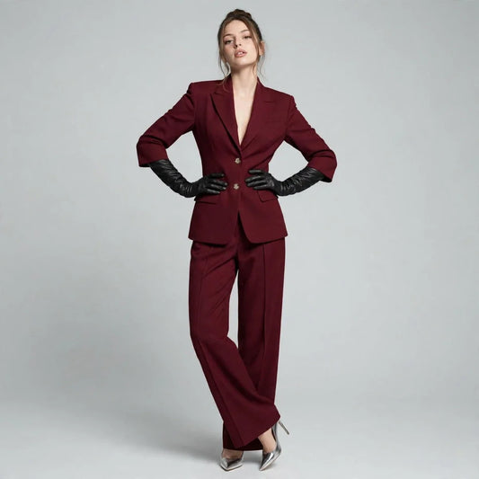 Woman with hands on hips in a burgundy tailored blazer and wide-leg pants set with black gloves, gray background.
