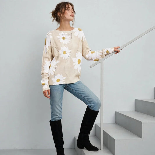 Model standing on stairs wearing a cream daisy-print knit sweater with blue jeans and black knee-high boots, white background