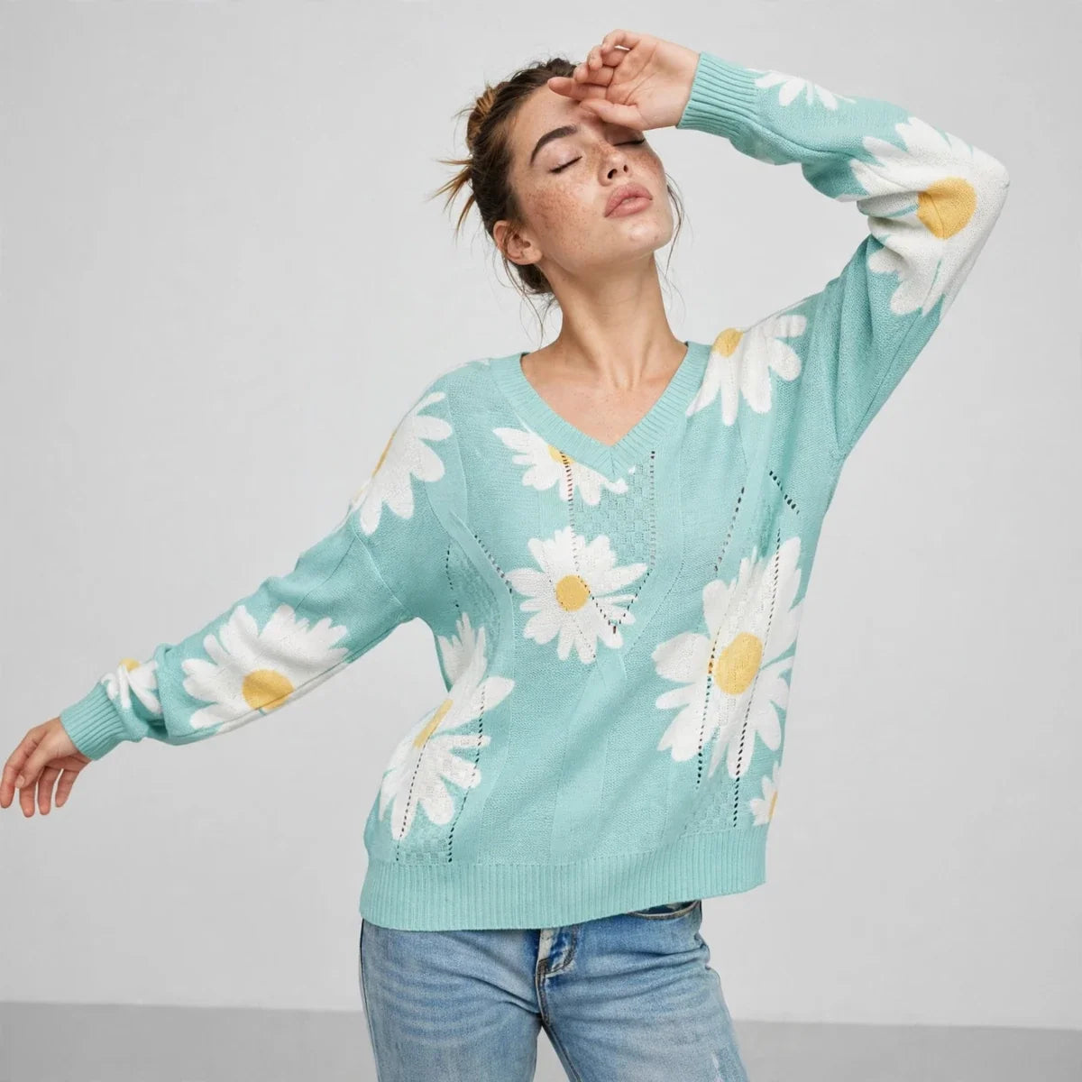 Model with arm raised wearing a mint green daisy-print knit sweater and blue jeans, front view on light background