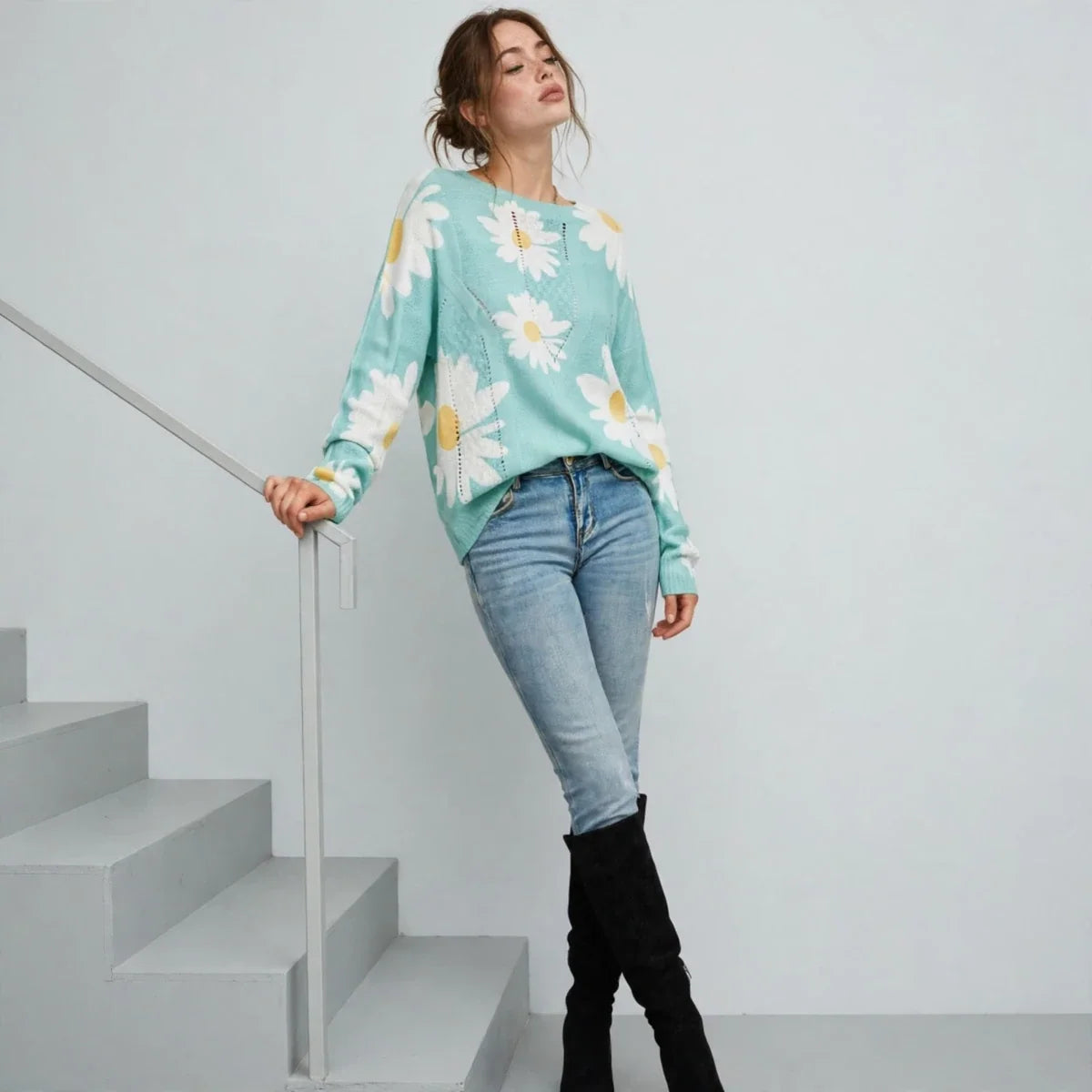 Model standing on stairs wearing a mint green daisy-print knit sweater with blue jeans and black boots, white background