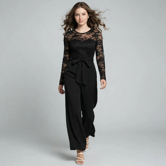 Model walking toward camera in a black lace long-sleeve jumpsuit with sheer lace bodice and tied waist sash, studio.