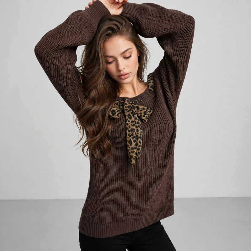 Woman with arms raised wearing a brown knit sweater with a leopard-print bow at the neckline, black pants, gray studio.