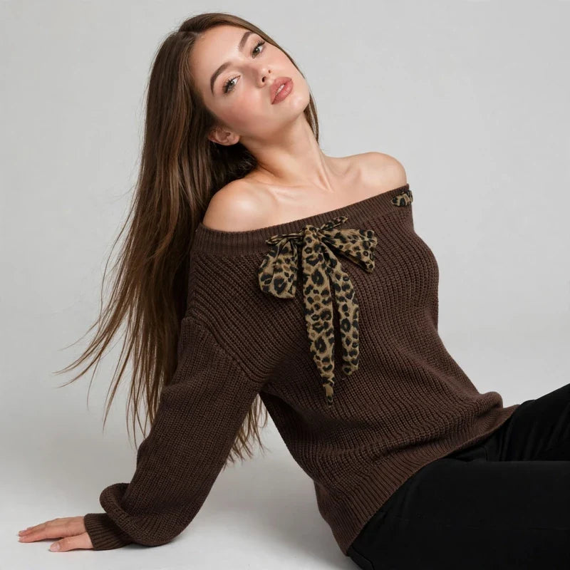 Woman reclining in a brown off-shoulder knit sweater with a leopard-print bow at the neckline, black pants, gray studio.
