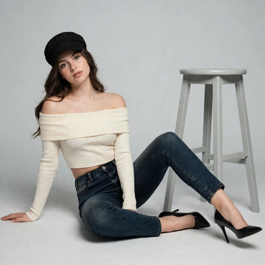 Woman seated on the floor wearing a cream off-shoulder ribbed knit sweater, dark jeans, black beret and heels, gray studio.