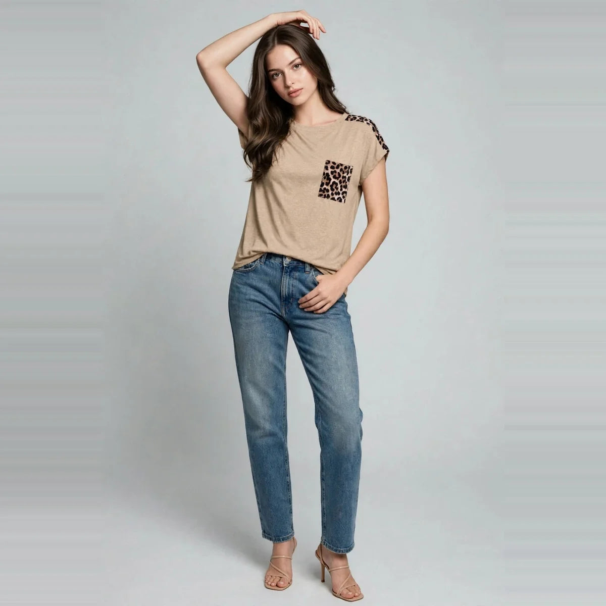 Model in a beige cap-sleeve tee with leopard print pocket and blue jeans, hand on head, barefoot, gray studio background.