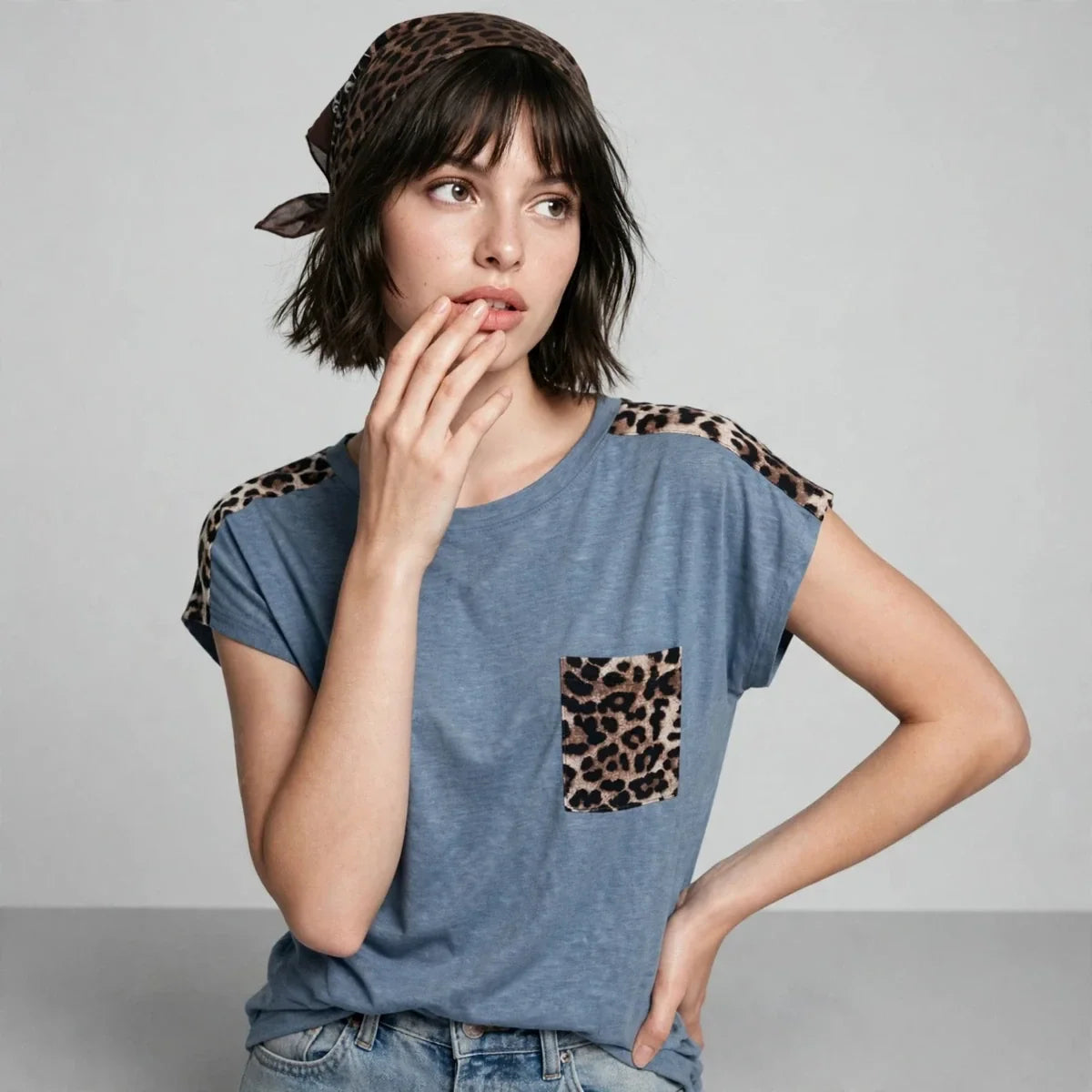 Close-up of a model in a blue cap-sleeve tee with leopard print shoulders and pocket, wearing a headscarf, gray background.