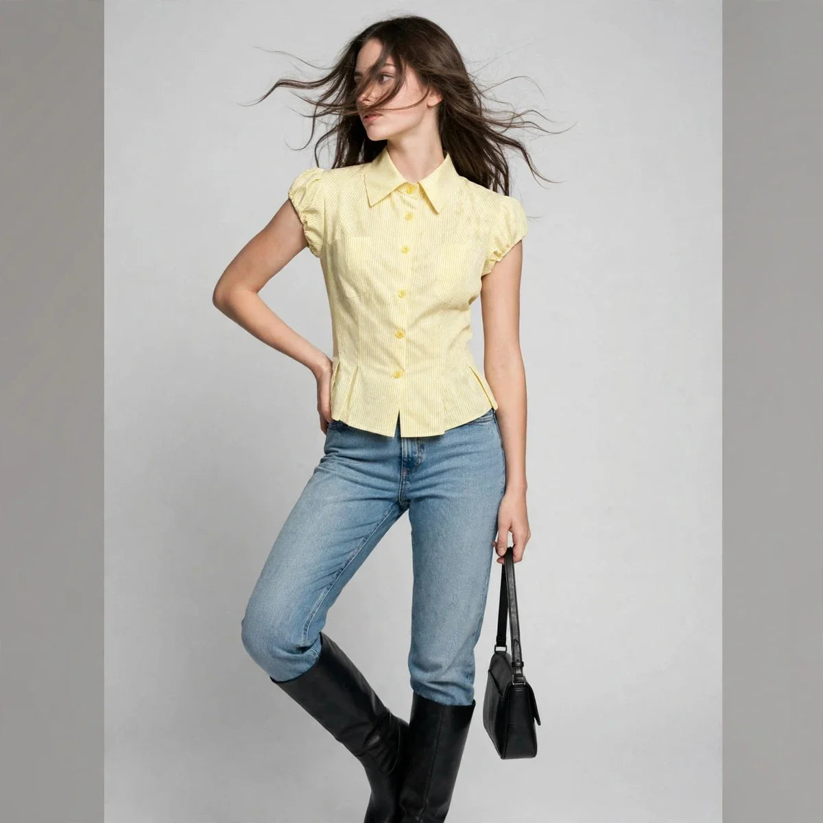 Full-body view of a woman in a yellow pinstripe puff-sleeve shirt, blue jeans, black boots, holding a handbag.