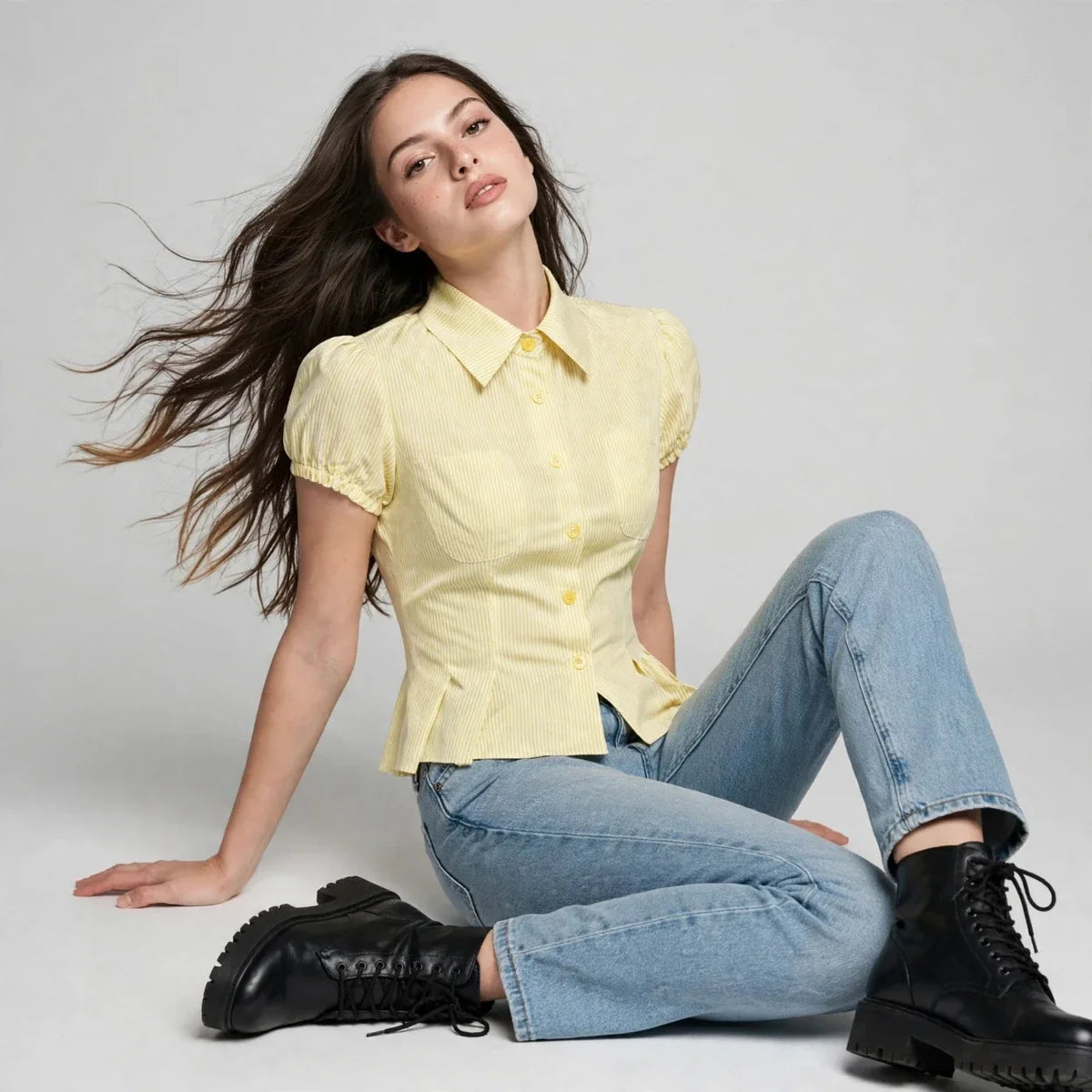 A woman seated on the floor wearing a yellow pinstripe puff-sleeve shirt, blue jeans, and black boots.