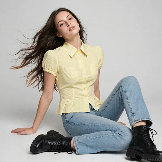 A woman seated on the floor wearing a yellow pinstripe puff-sleeve shirt, blue jeans, and black boots.