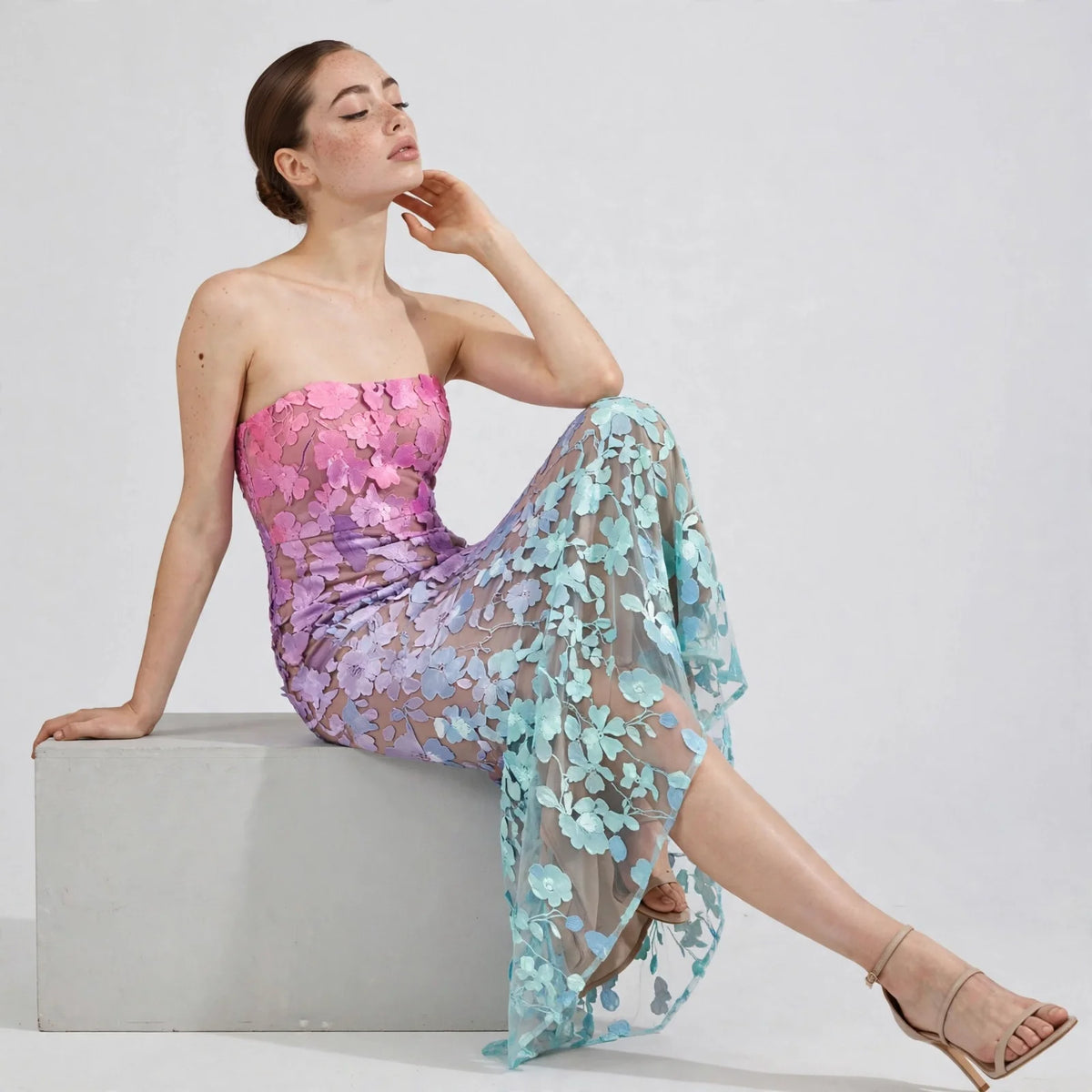 Woman seated on a block wearing a pink-to-mint ombré floral strapless maxi dress and nude heels, white studio background.