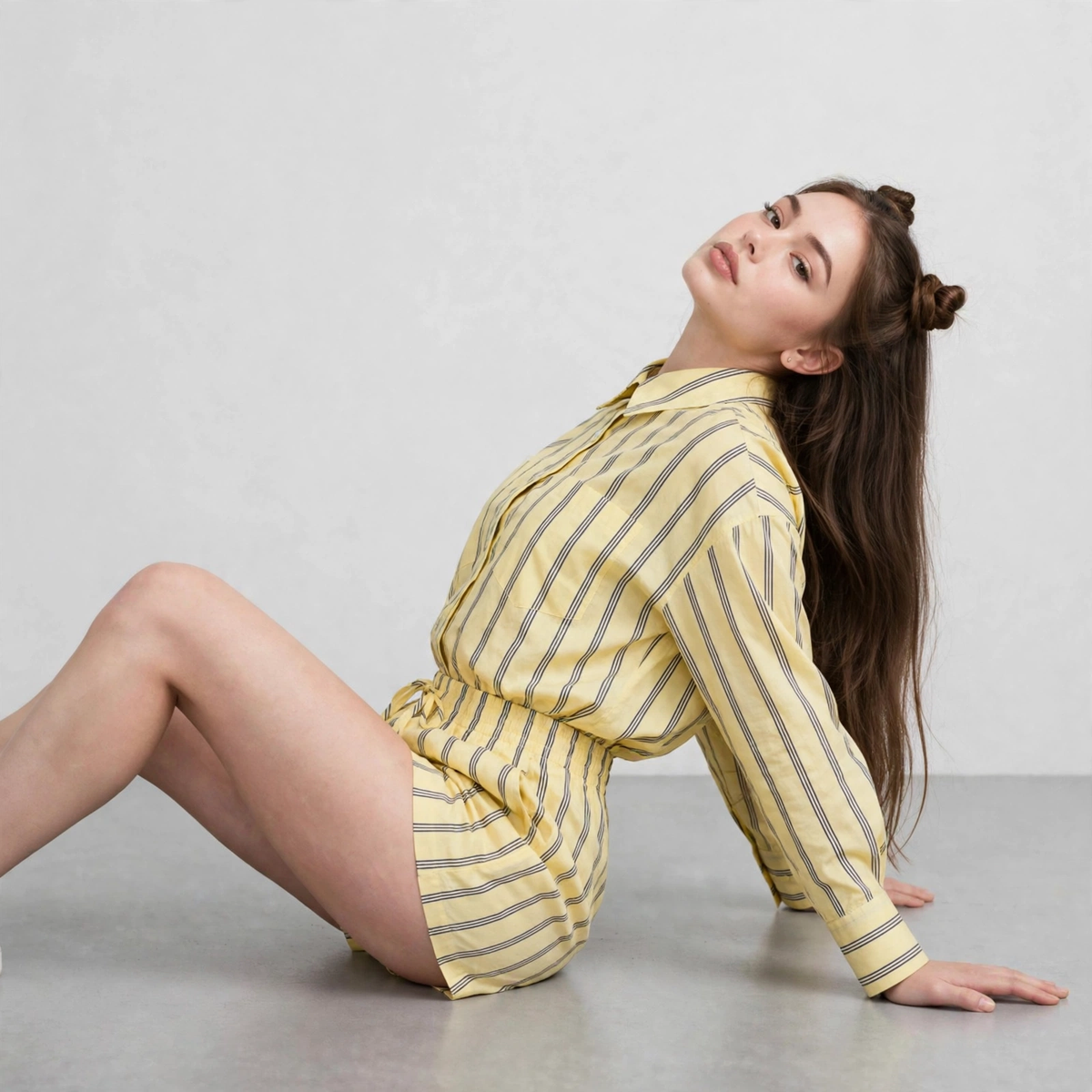Woman sitting on floor in yellow and grey striped button-up shirt and shorts set, leaning back, grey studio backdrop.