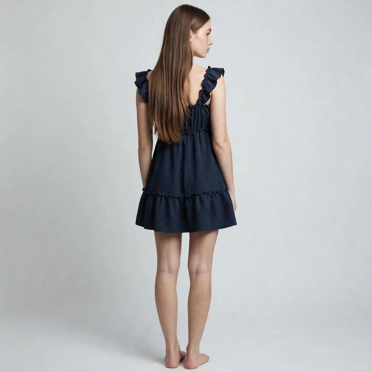 Back view of woman in navy ruffled square-neck mini dress, ruffle straps, tiered skirt, barefoot, grey backdrop.