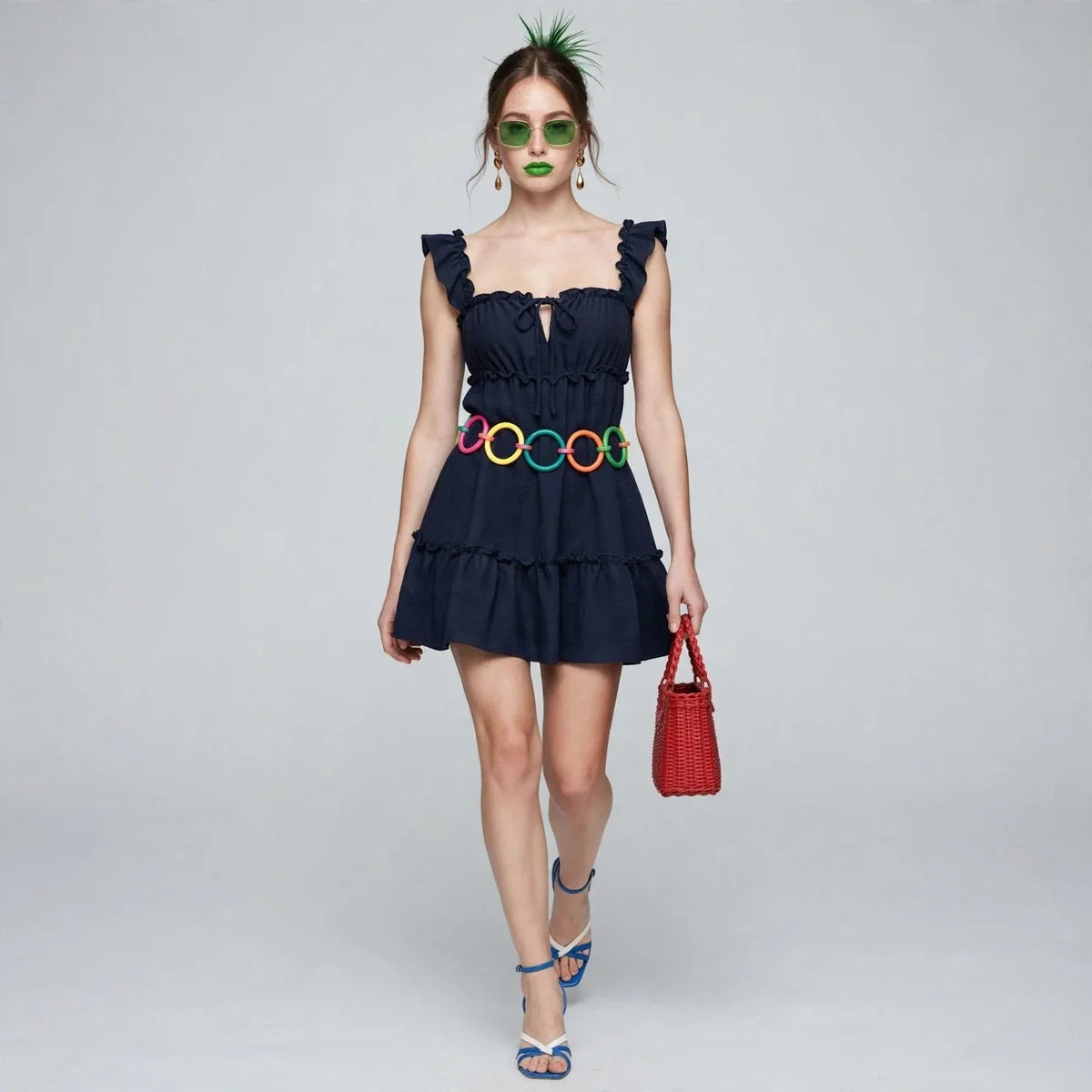 Woman in navy ruffled square-neck mini dress with colorful belt, red mini bag and blue heels, front view, grey backdrop.