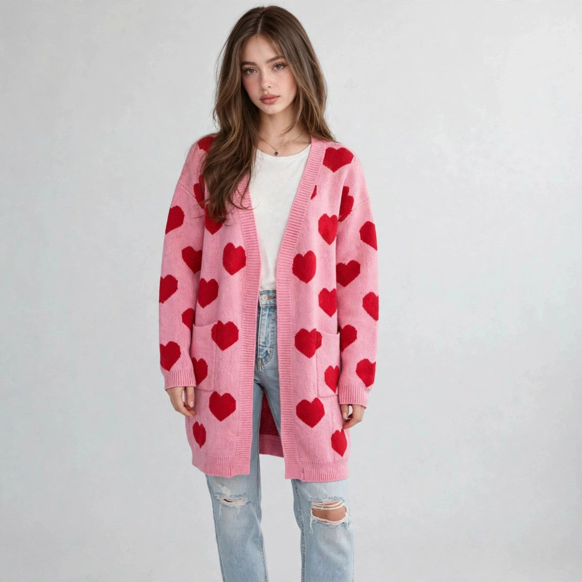 Model standing in a pink open-front cardigan with red hearts over a white top and ripped jeans, gray background.