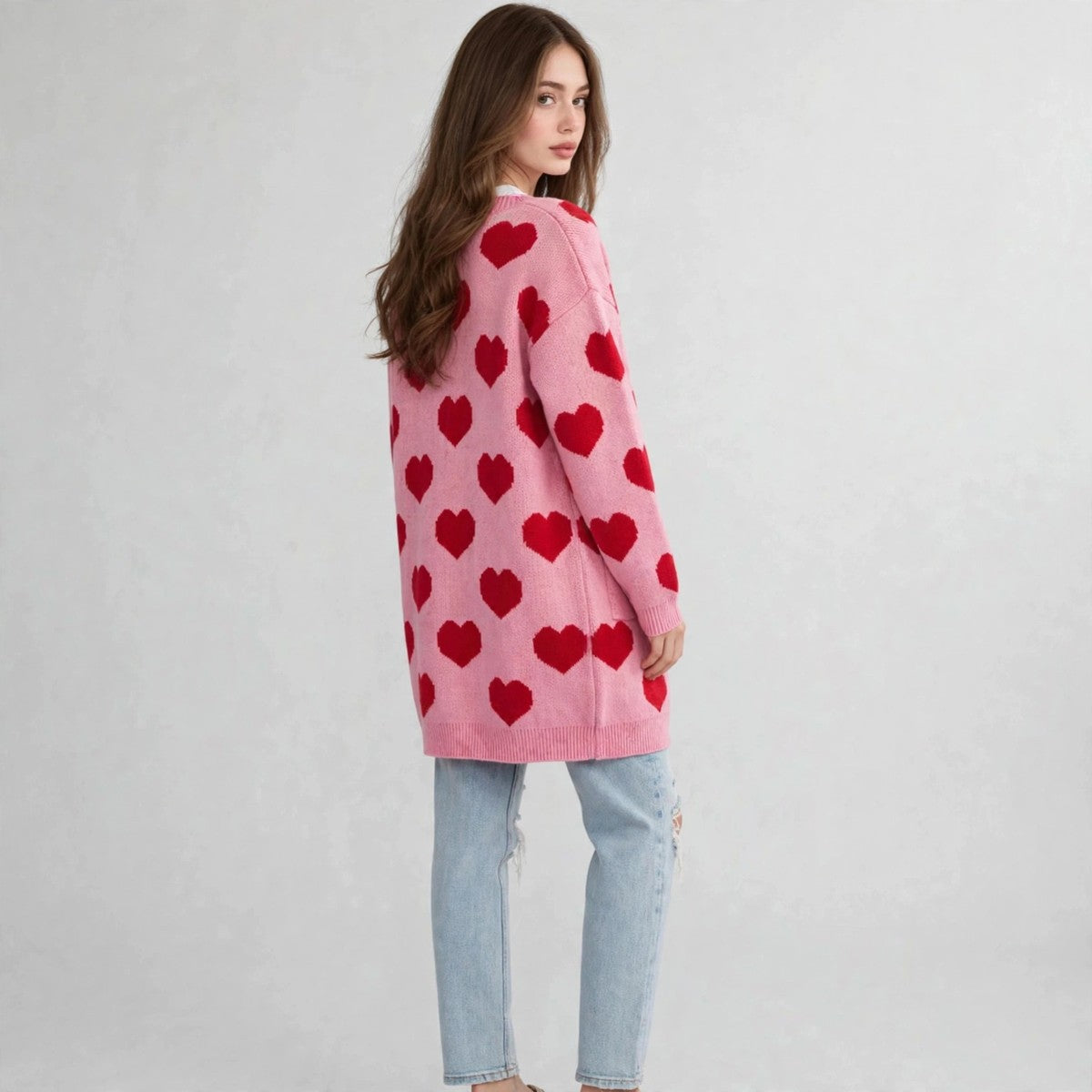 Back view of a model in a pink open-front cardigan with red heart print and light-wash ripped jeans, gray studio background.