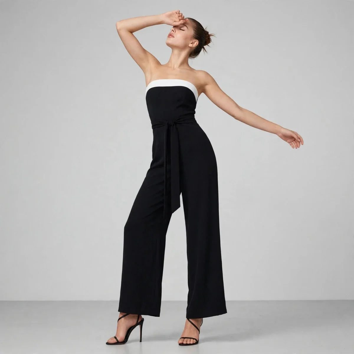 Front view of a black strapless wide-leg jumpsuit with white contrast bust band, model with arm raised, gray studio.