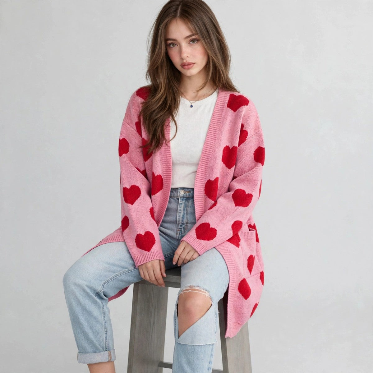 Model seated on a stool in a pink open-front cardigan with red hearts over a white top and ripped jeans, gray background.