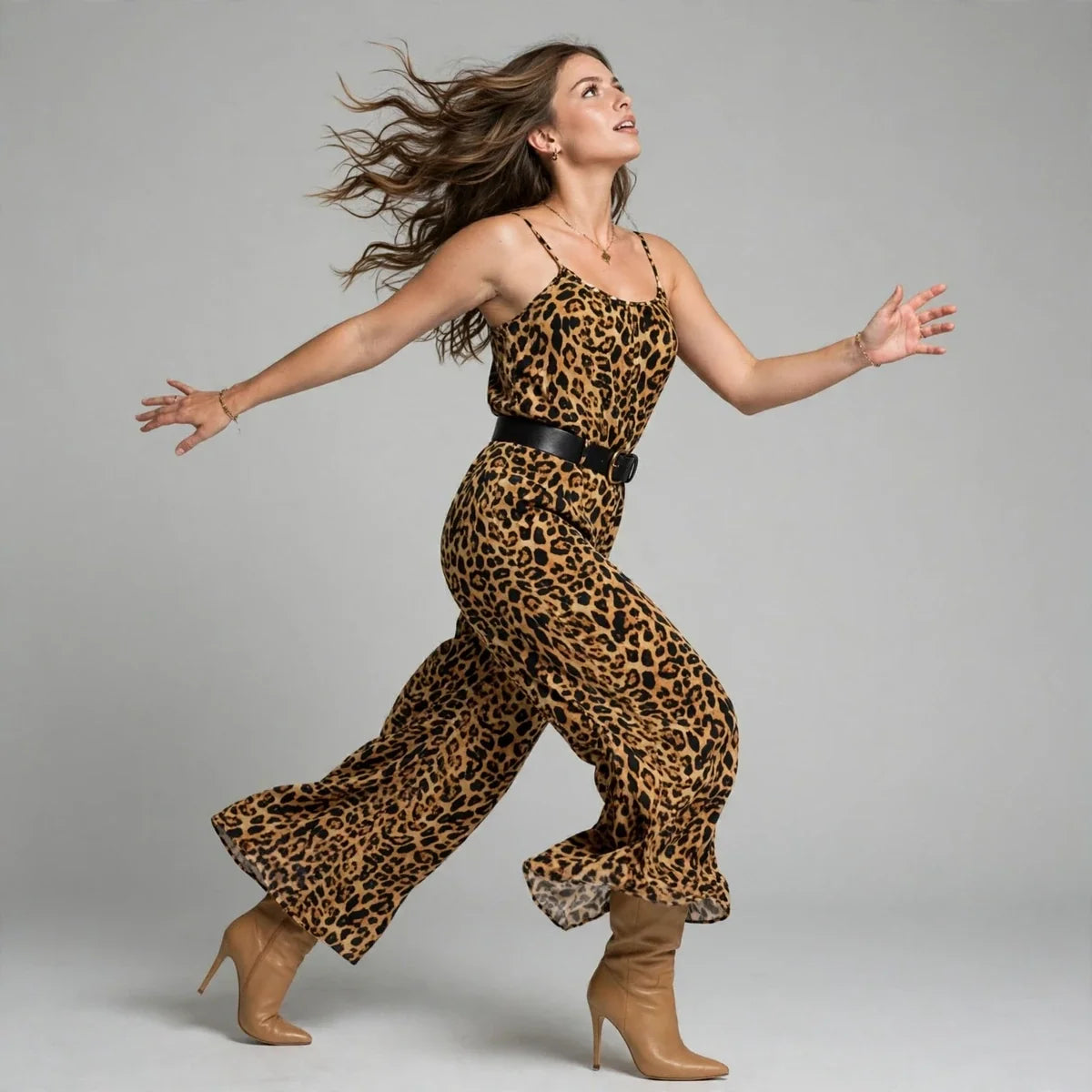Woman mid-dance wearing a leopard print wide-leg jumpsuit with scoop neckline and tan boots, gray studio background.
