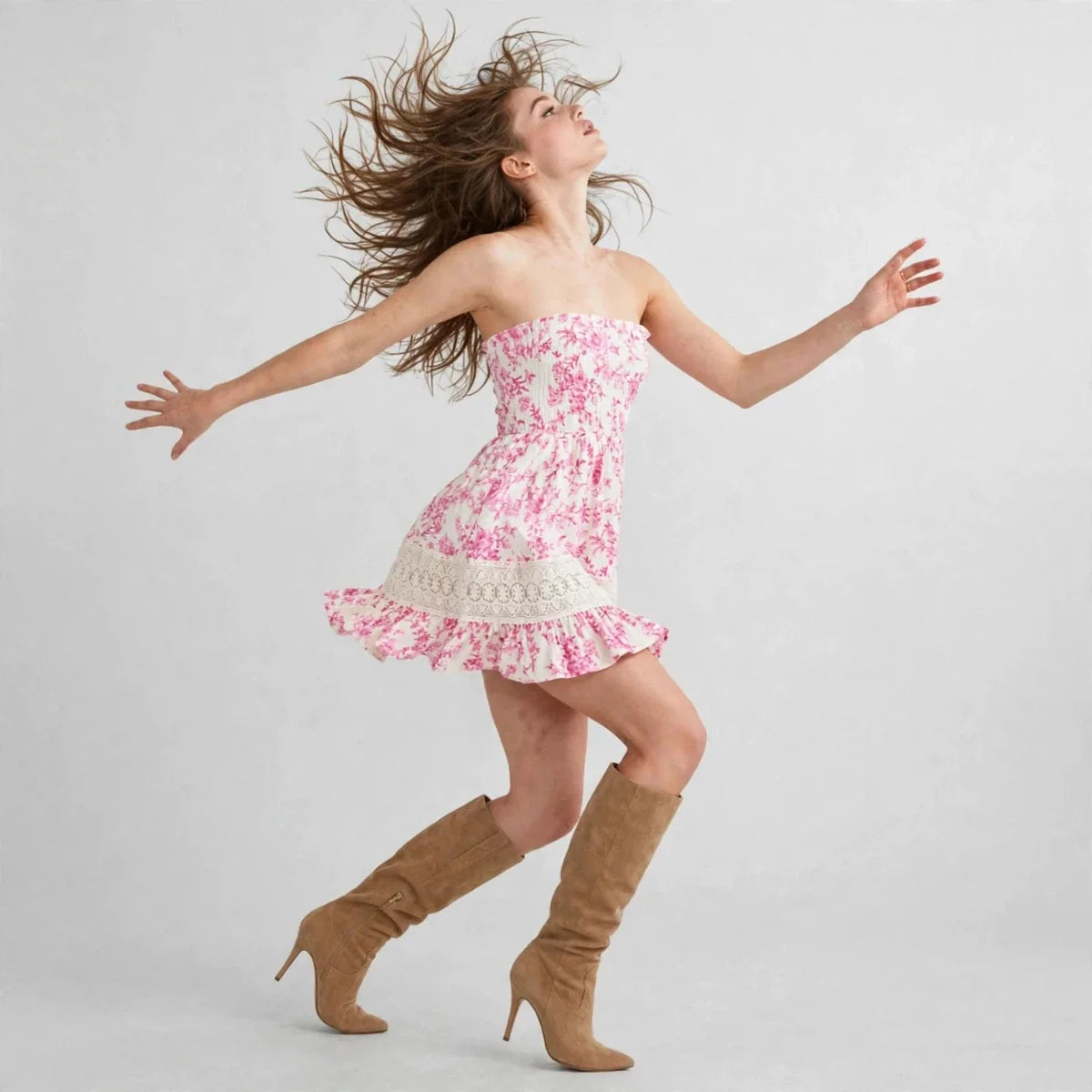 Woman jumping in a pink floral strapless mini dress with a ruffled hem, wearing tan knee-high boots, gray background.