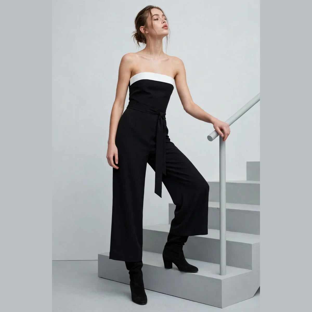 Model posing on studio stairs in a black strapless tube jumpsuit with white contrast bust band and black ankle boots.