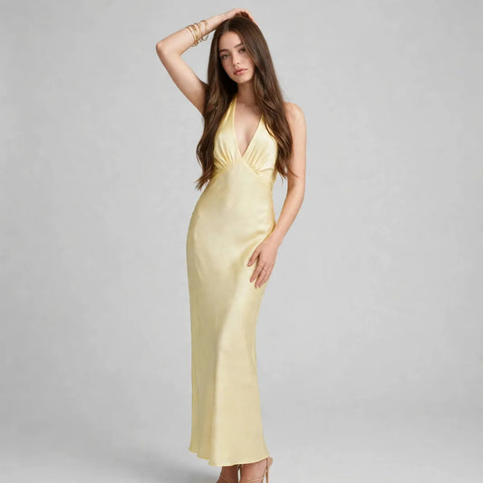 Woman with hand in hair in pale yellow satin halter-neck maxi dress, plunging neckline, front view, grey studio backdrop.